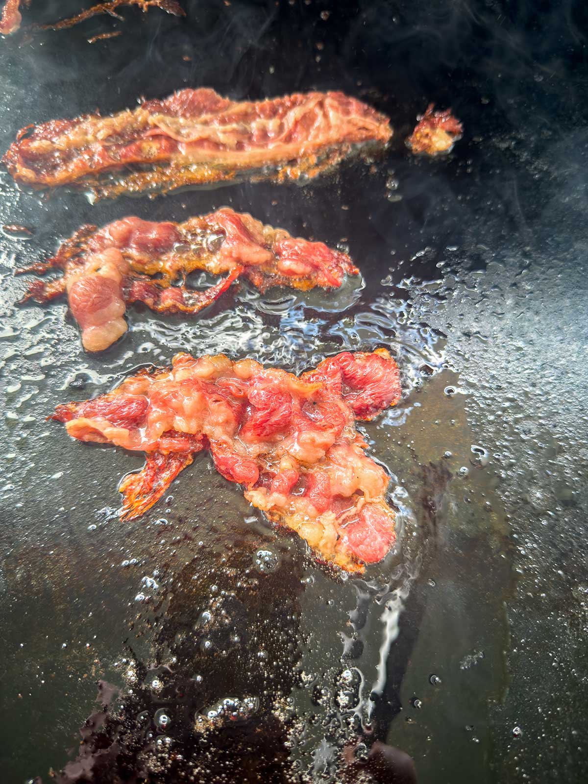 Crispy bacon strips sizzling on a greasy, black stovetop griddle with bubbles of hot oil surrounding the meat.