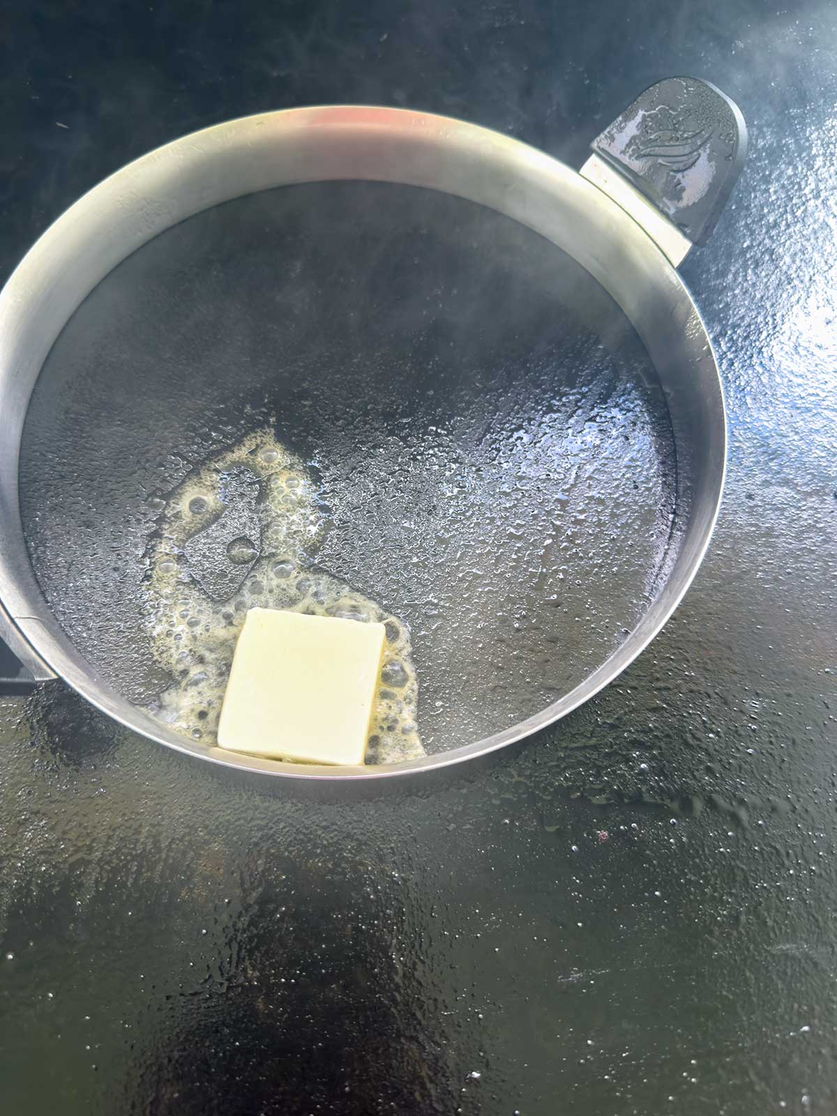 A metal ring mold on a dark griddle with a melting pat of butter inside. Steam rises as the butter sizzles and begins to bubble around the edges.