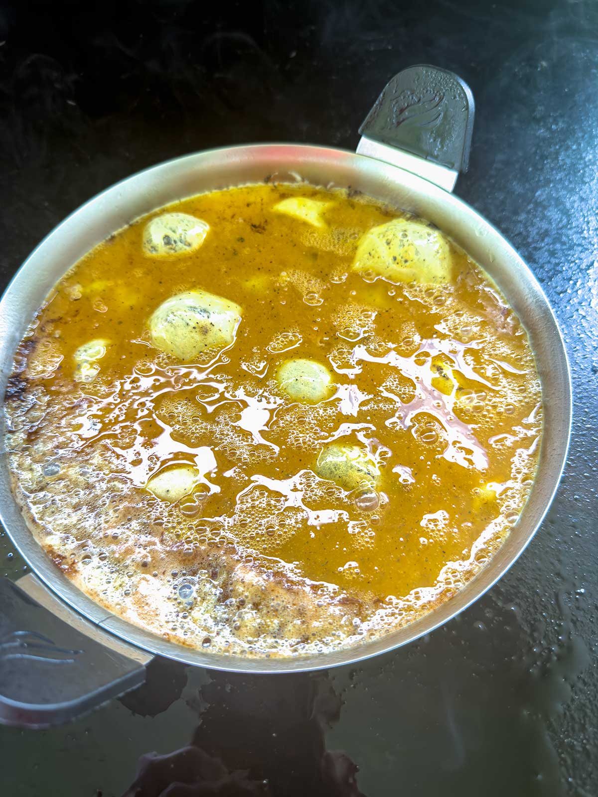 A metal pot filled with bubbling yellow-orange soup or curry with round dumplings or vegetables, sitting on a dark surface. Steam rises from the hot, freshly cooked dish.