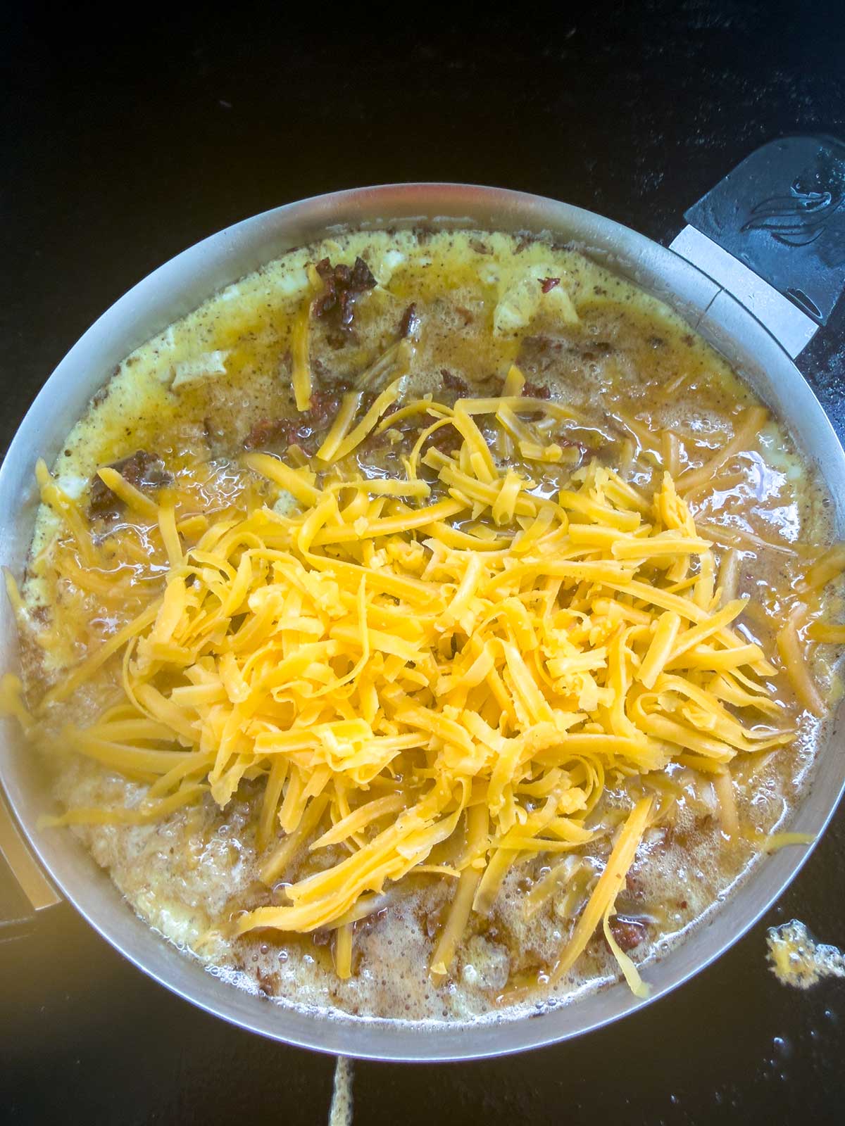 A frying pan filled with a partially cooked omelet topped with a generous amount of shredded cheddar cheese, sitting on a dark stovetop.