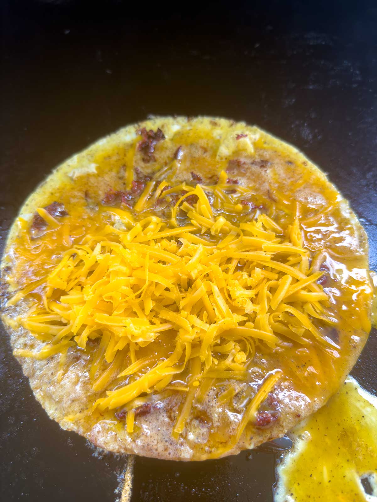 A close-up of an egg omelette cooking on a dark skillet, topped with shredded cheddar cheese and bits of cooked bacon or sausage.