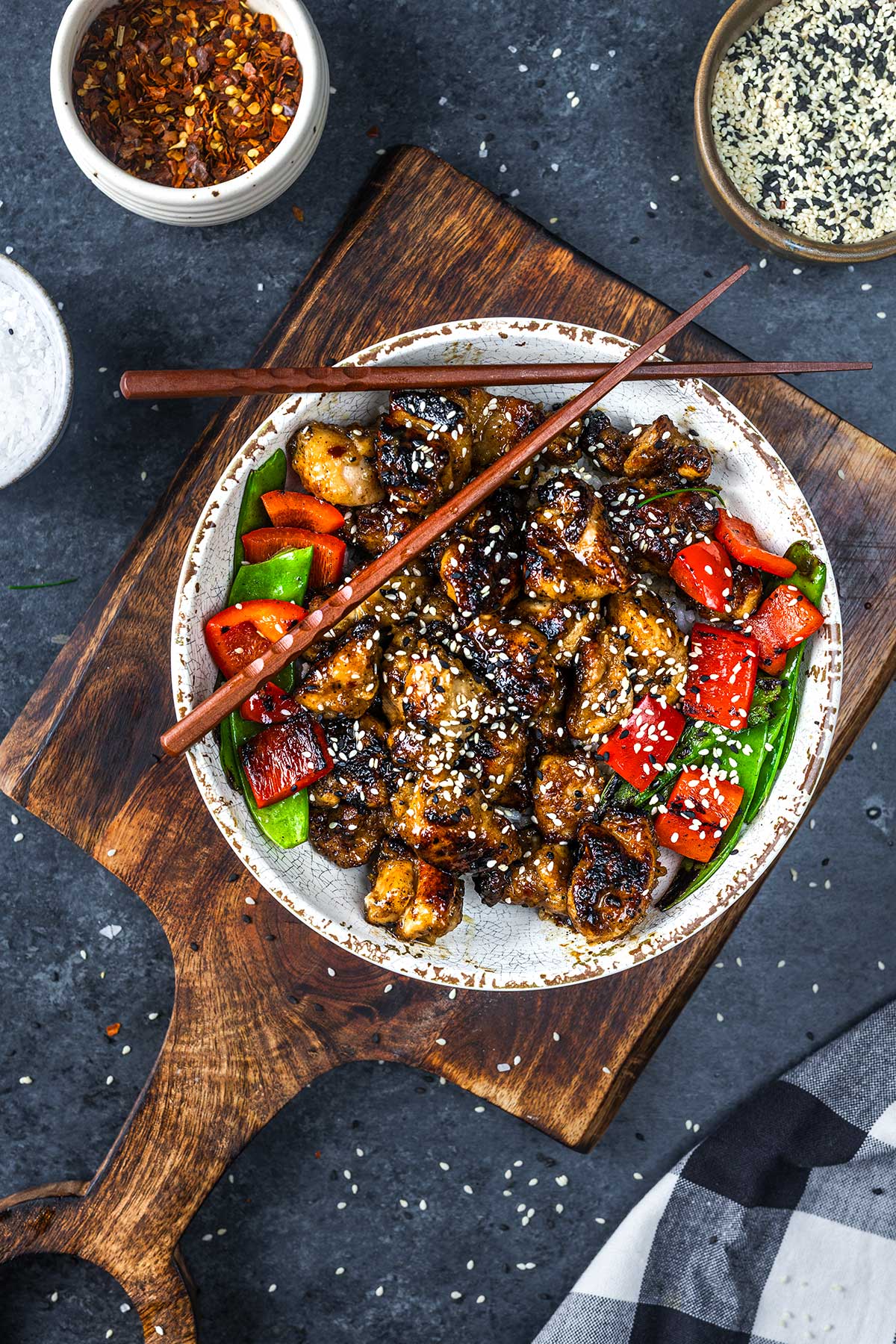 A bowl of stir-fried chicken with red bell peppers and green vegetables, topped with sesame seeds. Wooden chopsticks rest on the bowl, which is placed on a wooden board. Small bowls of spices and sesame seeds are nearby.