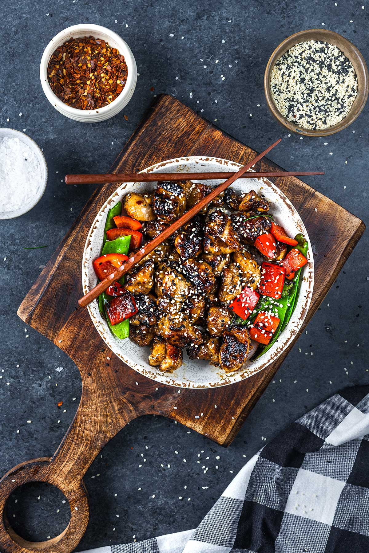 A bowl of Blacstone Orange Chicken with red bell peppers and green vegetables, topped with sesame seeds. Chopsticks rest on the bowl, which is set on a wooden board, with bowls of spices and a black-and-white cloth nearby.