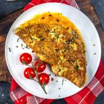 A golden-brown omelette garnished with chives and chili flakes is served on a white plate with a small bunch of cherry tomatoes. The plate sits on a red checkered napkin and a wooden board.