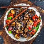 A bowl of stir-fried chicken glazed in sauce, garnished with sesame seeds, served with red bell peppers and snap peas. Wooden chopsticks rest on the bowl, set on a rustic wooden board.