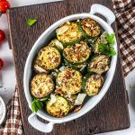 A white oval dish filled with grilled zucchini slices, garnished with herbs, grated cheese, and small pieces of nuts, sits on a wooden board next to a checkered napkin and scattered cherry tomatoes.