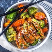 A bowl of rice topped with glazed chicken slices, steamed broccoli, and carrots, garnished with sesame seeds and green onions. Black chopsticks rest on the bowl, which sits on a striped napkin.