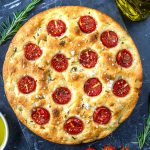 A round focaccia bread topped with halved cherry tomatoes, rosemary, and flaky sea salt, displayed on a dark surface with olive oil and rosemary sprigs nearby.