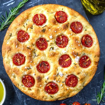 A round focaccia bread topped with halved cherry tomatoes, rosemary, and flaky sea salt, displayed on a dark surface with olive oil and rosemary sprigs nearby.
