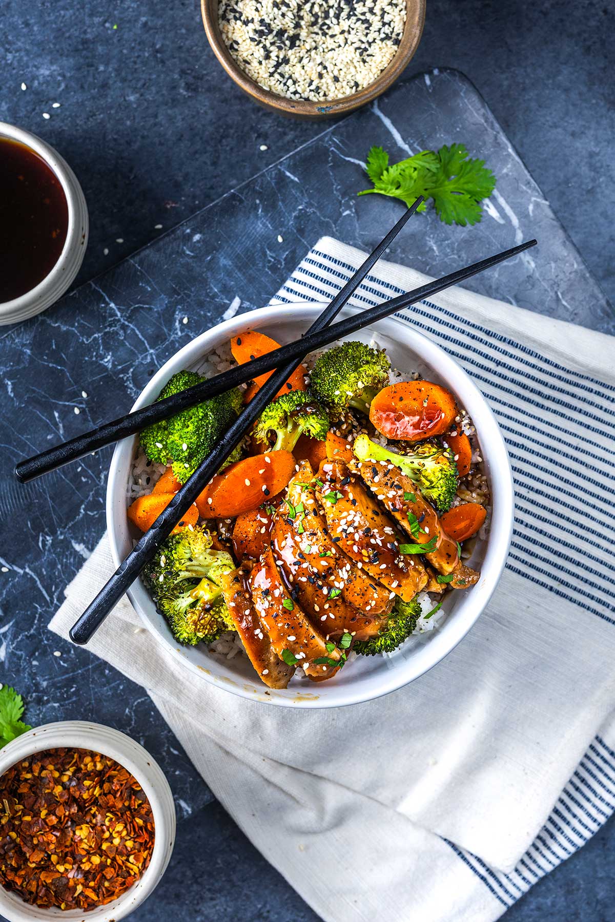 A bowl of stir-fried vegetables and sliced tofu topped with sesame seeds, with black chopsticks resting on top. Surrounding the bowl are small dishes of sauce, sesame seeds, and red pepper flakes on a dark surface.