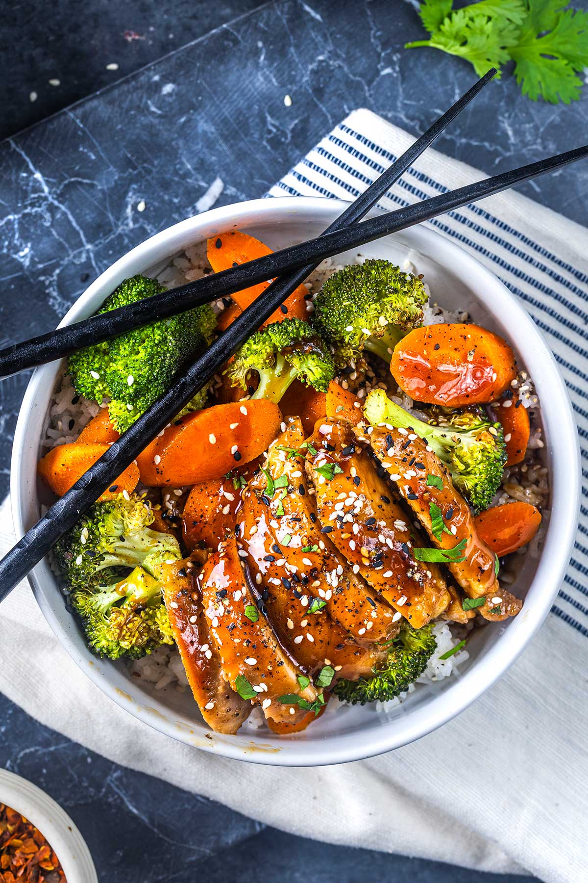 A bowl of rice topped with glazed sliced chicken, roasted broccoli, and carrots, sprinkled with sesame seeds, with a pair of black chopsticks resting on top.