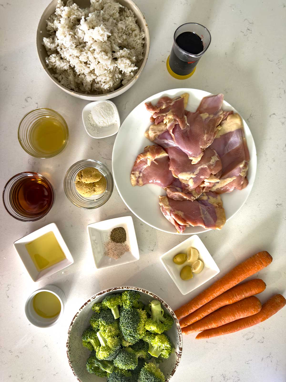Top-down view of ingredients for a teriyaki chicken bowl on a white surface, including bowls of rice, broccoli, raw chicken thighs, carrots, seasonings, garlic, oil, soy sauce, vinegar, and brown sugar.