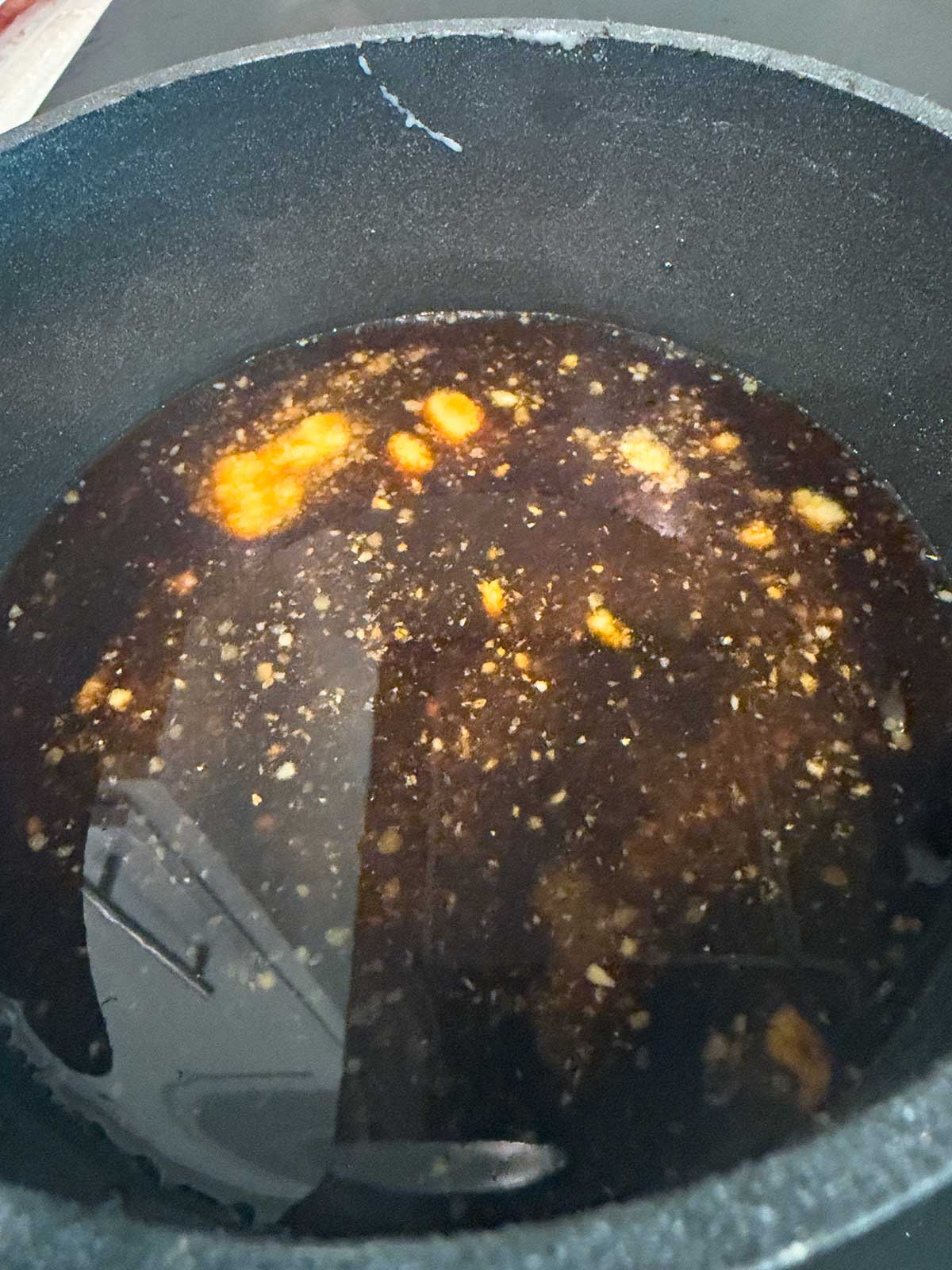 A close-up of a dark liquid with floating yellowish chunks and visible seasonings in a black cooking pot, capturing the simmering base for a savory teriyaki chicken bowl.
