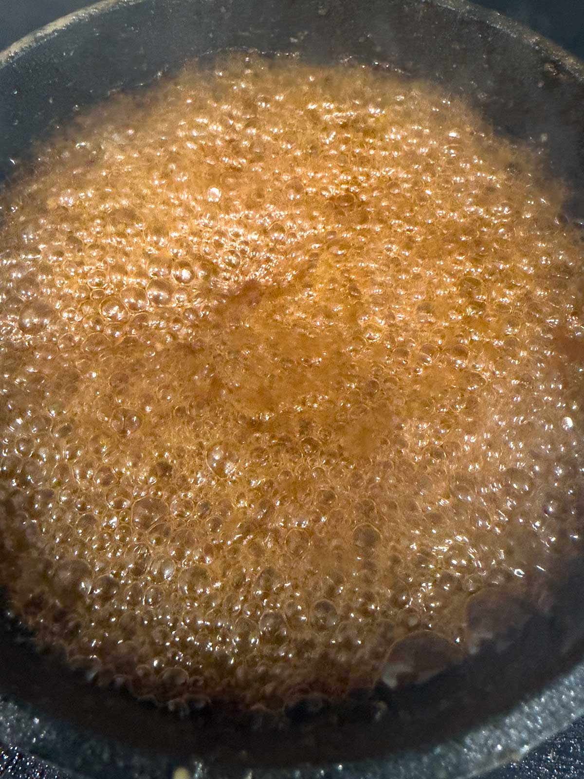 A close-up view of a pot with a bubbling, frothy brown teriyaki sauce in a dark pot.