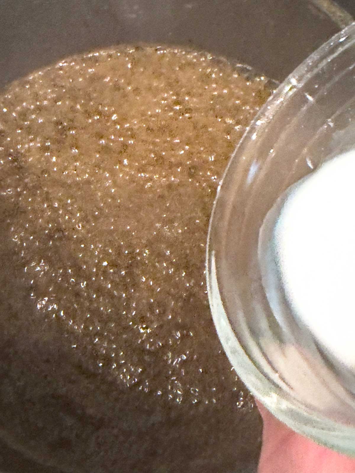 A cornstarch slurry being poured into boiling teriyaki sauce on the stove.