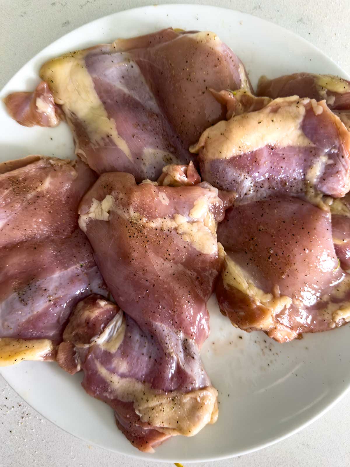 Raw, seasoned chicken thighs arranged on a white plate, ready for cooking—the perfect start for a flavorful teriyaki chicken bowl. The chicken is pink with visible seasoning and some fat, placed on a light-colored surface.