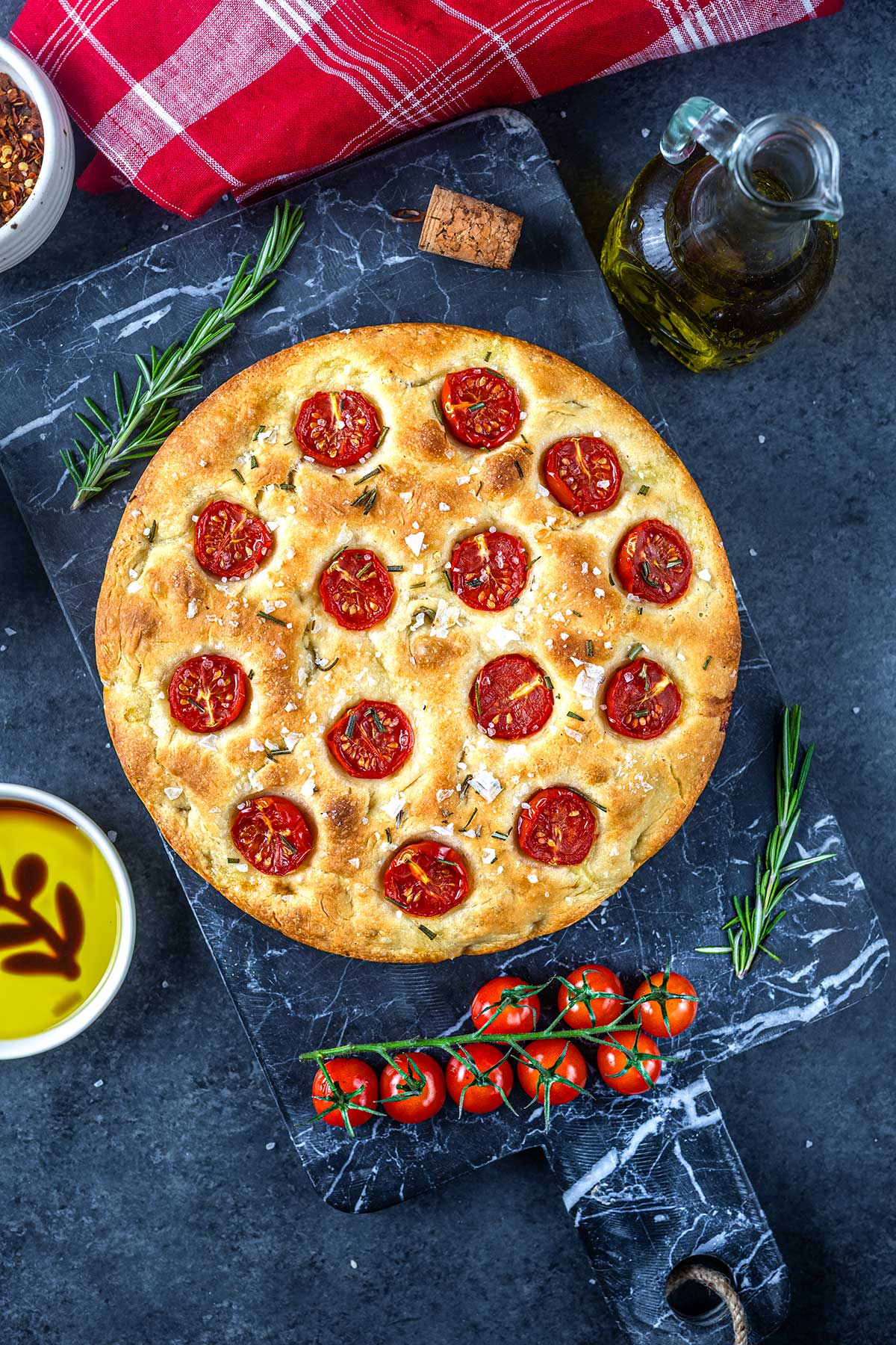 A round focaccia bread topped with halved cherry tomatoes and rosemary sits on a black marble board, surrounded by olive oil, fresh rosemary, cherry tomatoes on the vine, and a red checkered napkin.