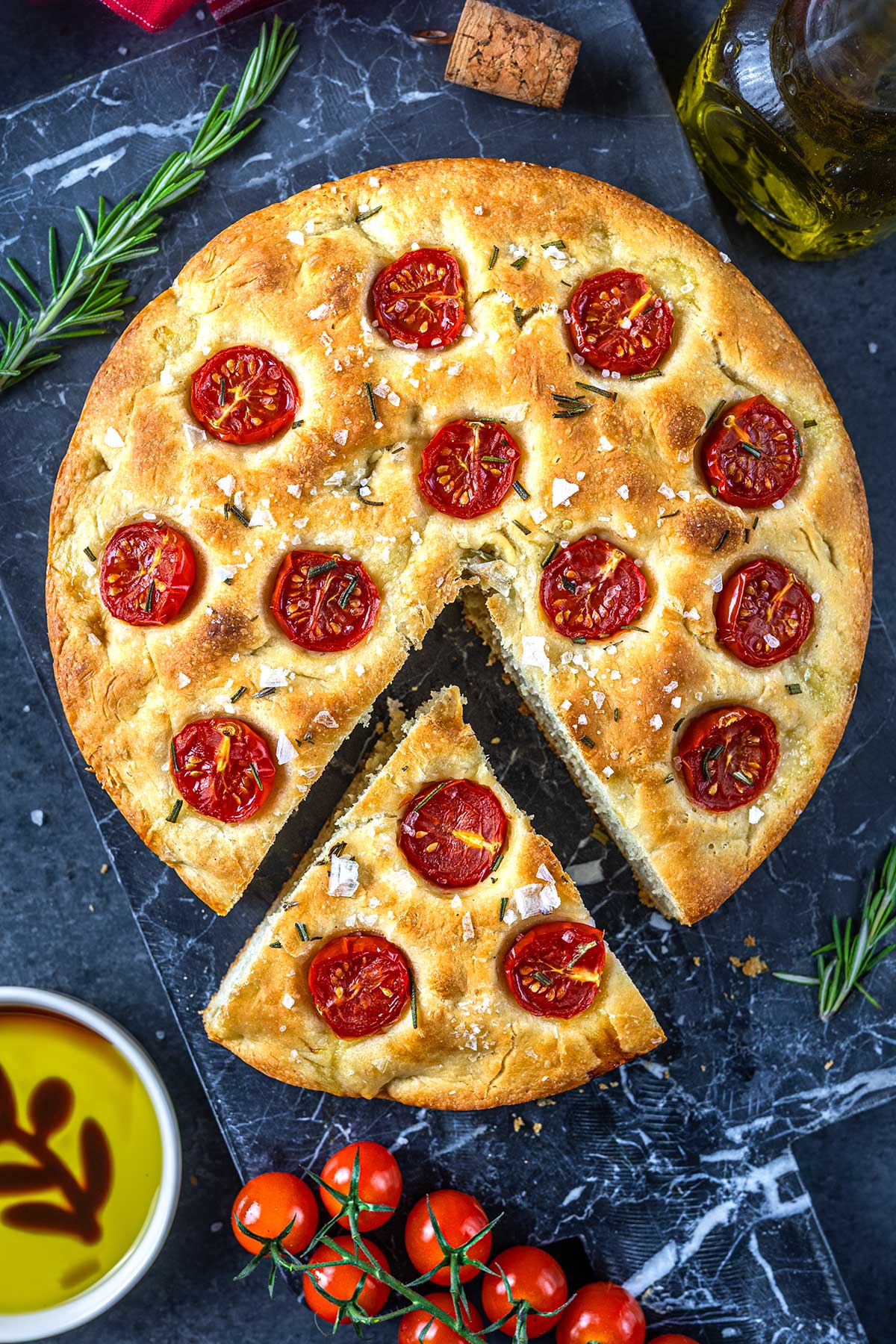 A round focaccia bread topped with cherry tomato slices, sea salt, and rosemary, with one triangular slice cut out. Surrounding it are fresh cherry tomatoes, rosemary sprigs, olive oil, and a cork on a dark surface.