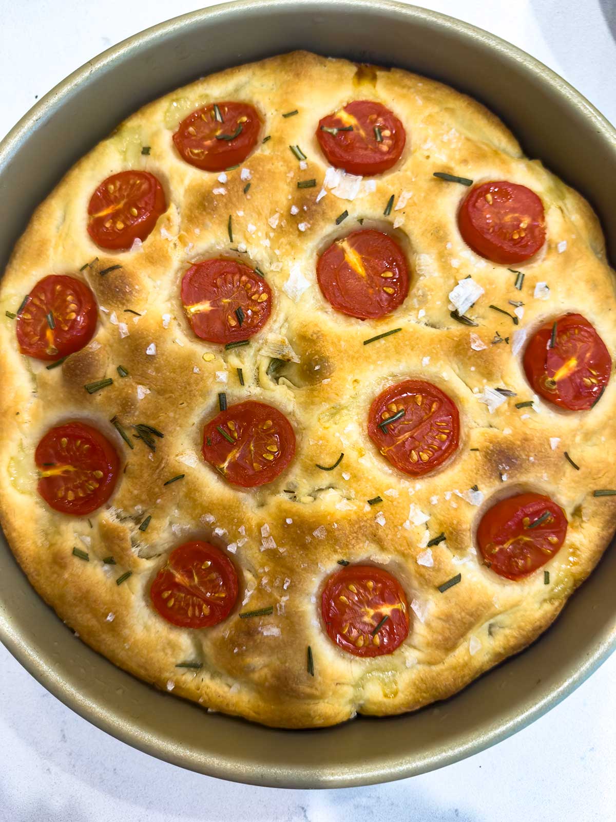 Round golden tomato focaccia bread in a baking pan, topped with halved cherry tomatoes, chopped herbs, and coarse sea salt.