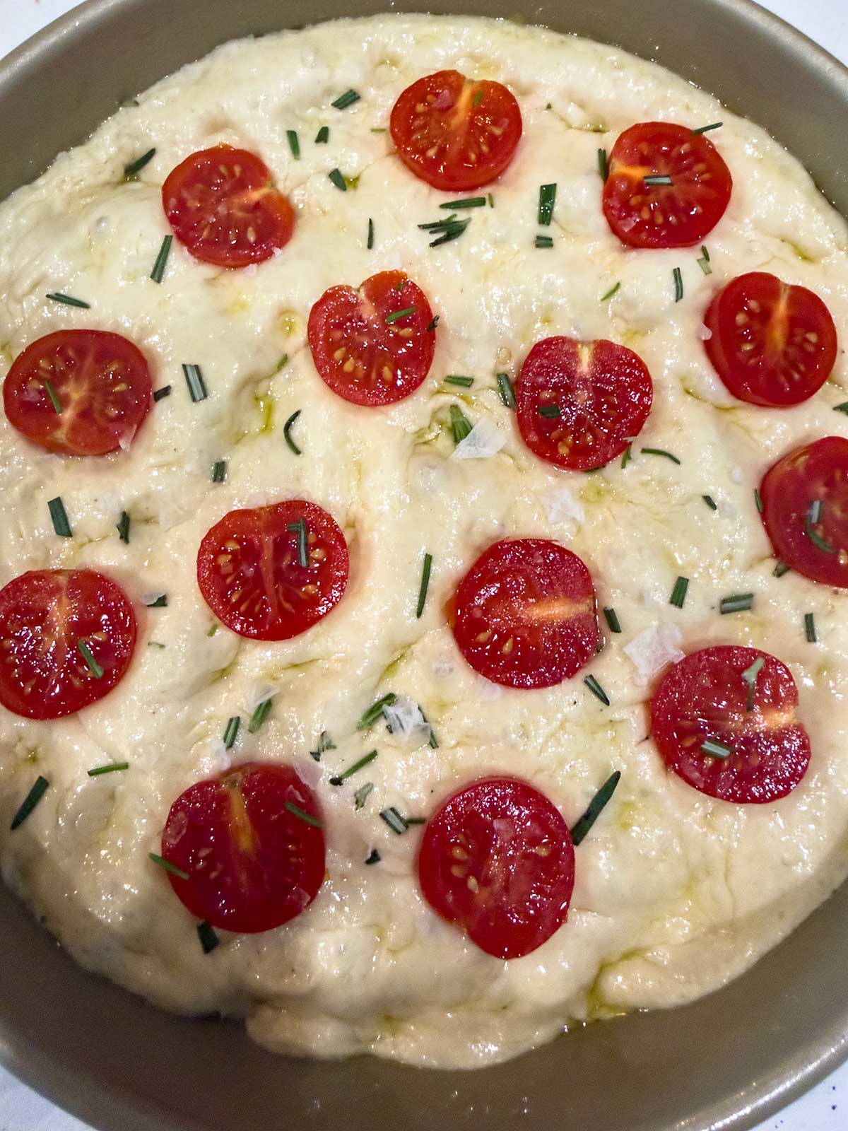 A round pan filled with tomato focaccia bread dough, topped with halved cherry tomatoes, chopped fresh rosemary, and a drizzle of olive oil, ready to be baked.