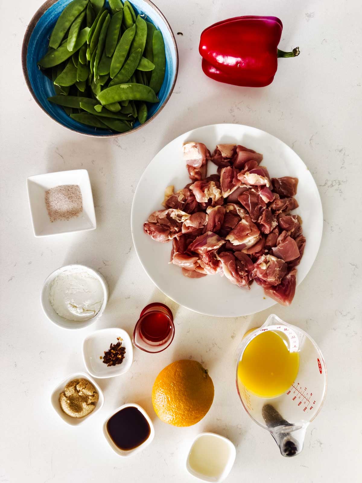 A white surface holds bowls and plates with snow peas, a red bell pepper, raw diced meat, seasonings, sauces, a whole orange, and a measuring cup of orange juice, arranged for meal preparation.