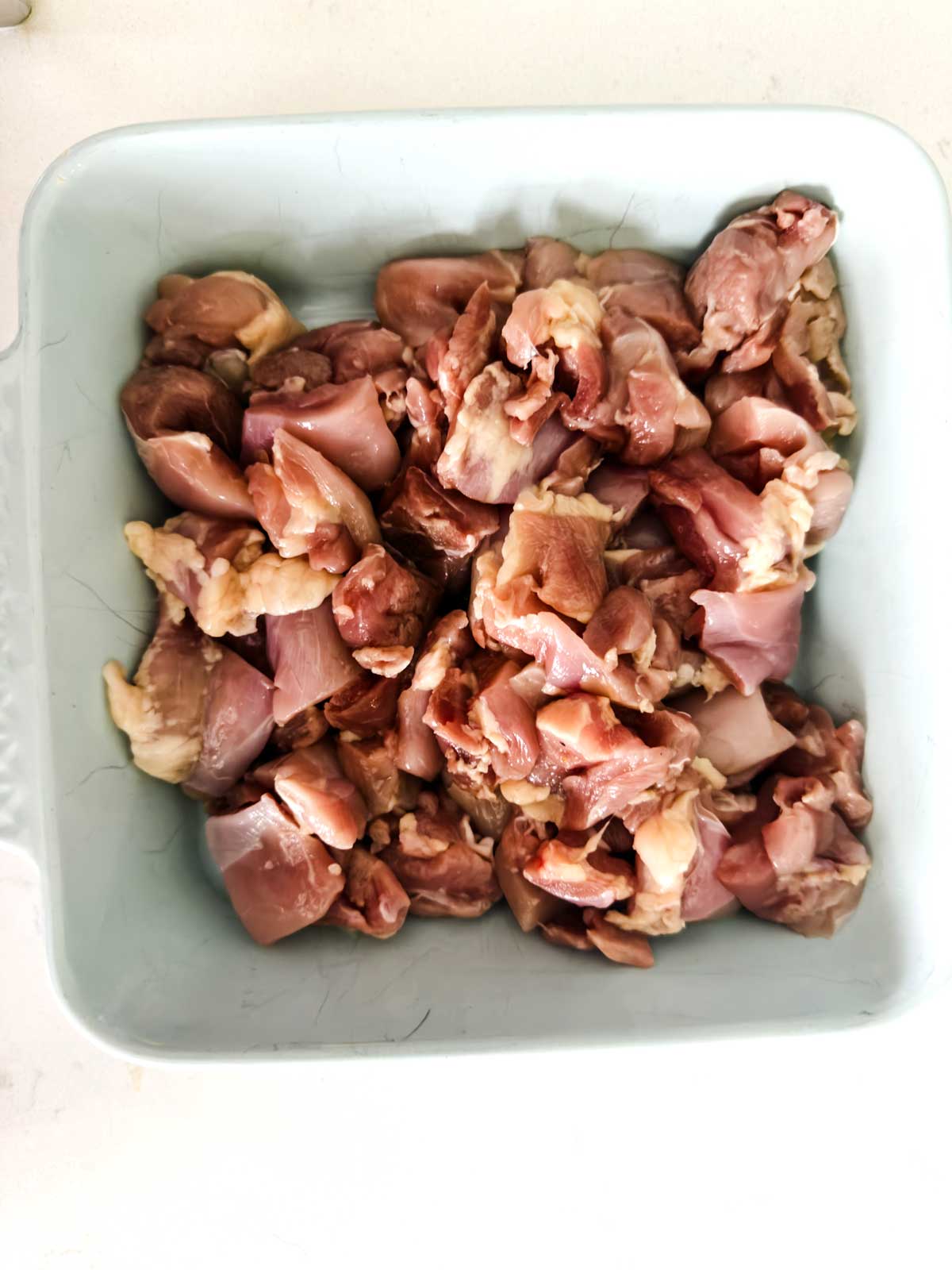 A light blue dish filled with raw, chopped pieces of meat, showing a mix of pink and light brown colors with visible fat. The dish is placed on a white surface.