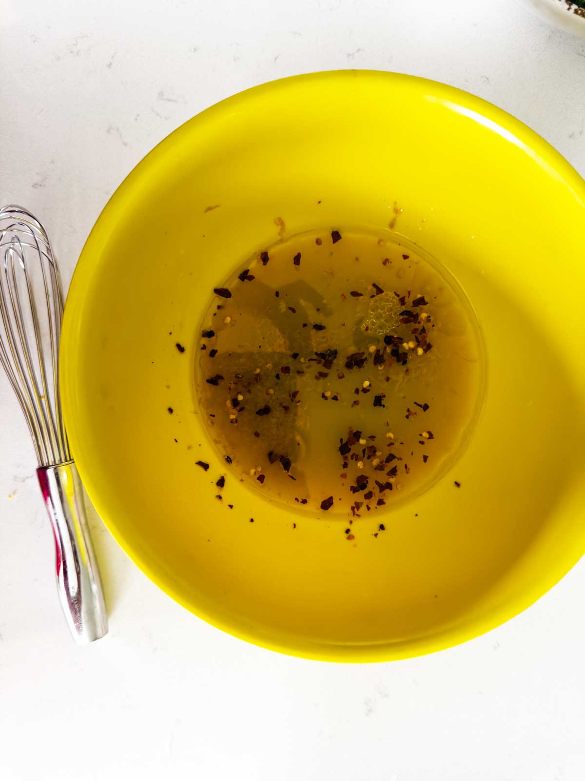 A metal whisk rests next to a bright yellow bowl containing a mixture of oil, seasonings, and red pepper flakes on a white surface.