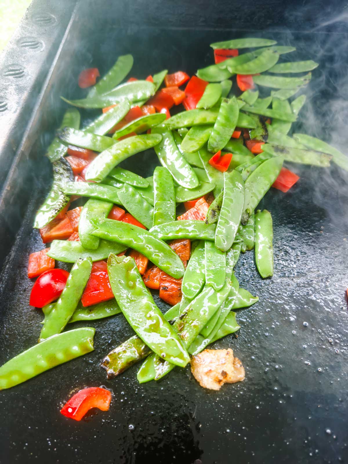 Snow peas and chopped red bell peppers sizzling on a hot griddle, with a bit of steam rising from the surface.