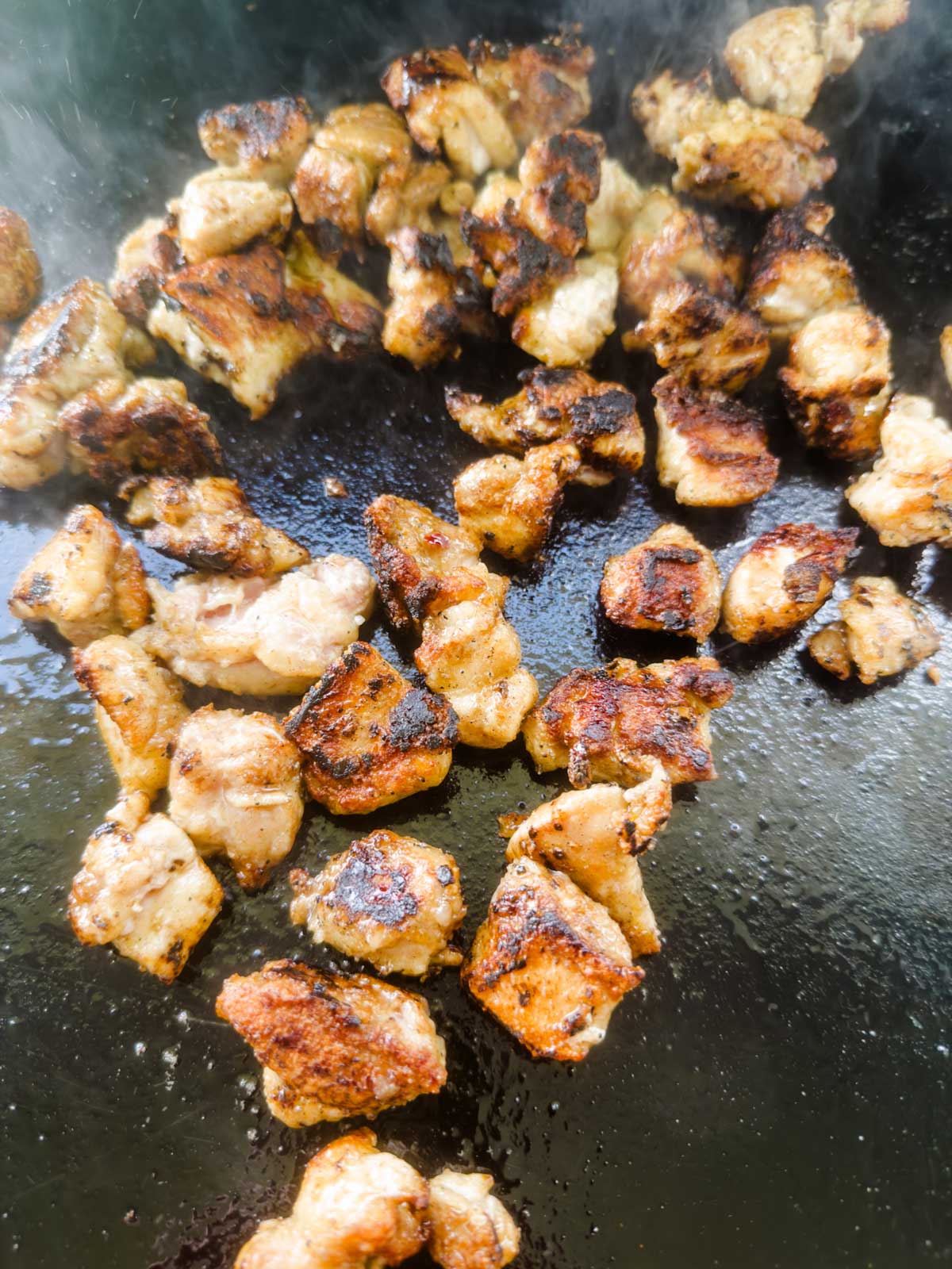 Small pieces of seasoned chicken are sizzling and browning on a hot, greased griddle, showing a crispy, golden-brown exterior with some steam rising.