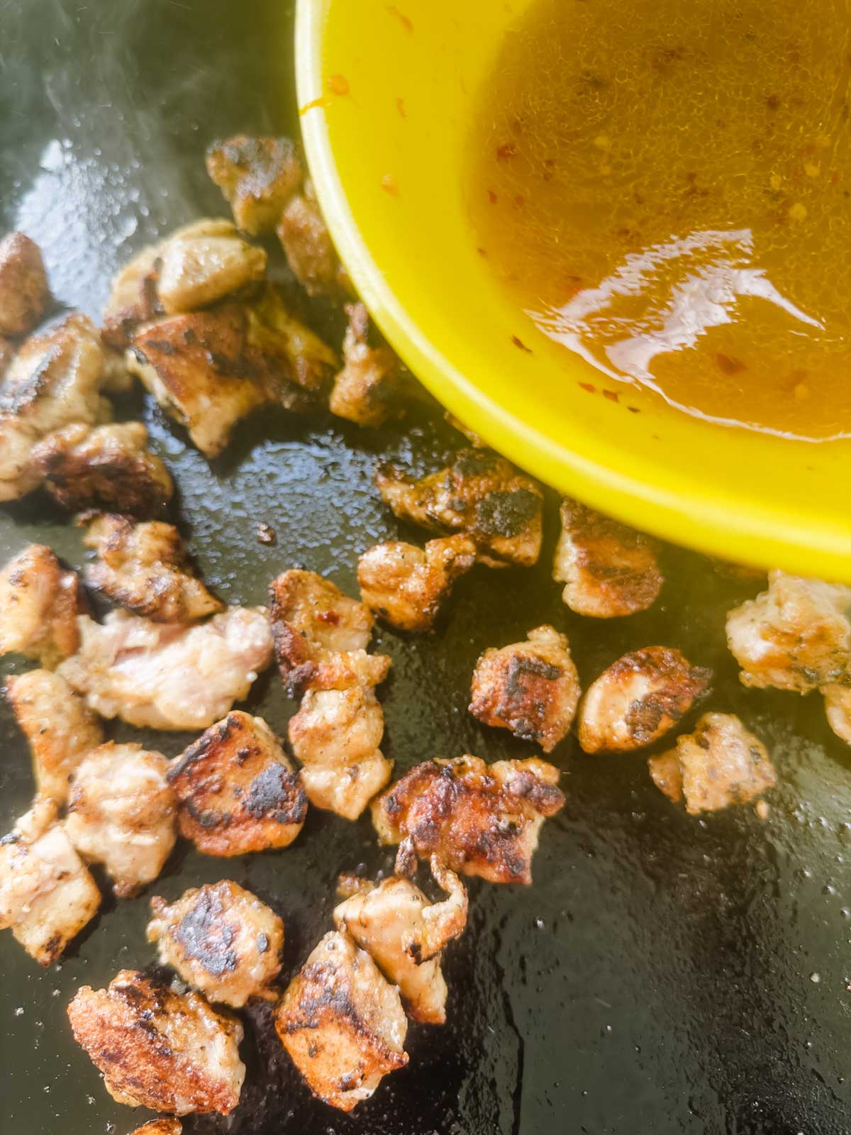 Close-up of grilled, browned pieces of meat on a hot, greasy surface, with a yellow bowl containing a reddish-brown sauce partially visible in the upper right corner.