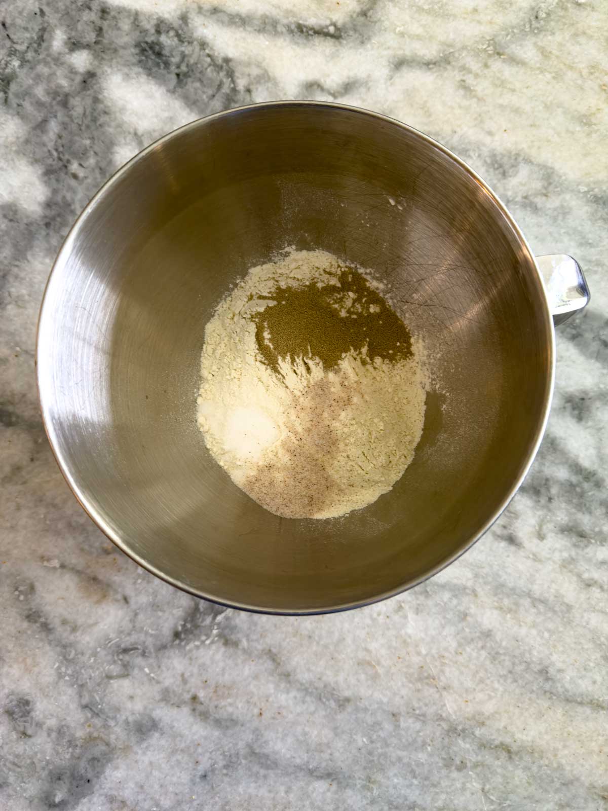 A metal mixing bowl containing flour and yeast for tomato focaccia bread sits on a gray marble countertop.