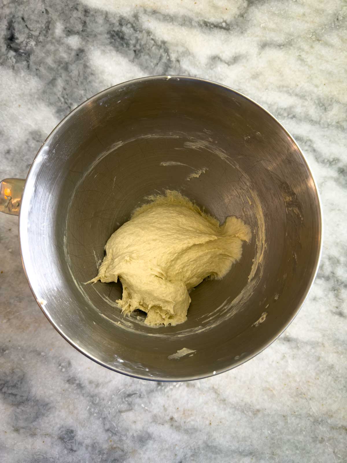 A ball of dough for tomato focaccia bread sits inside a stainless steel mixing bowl on a gray marble countertop.