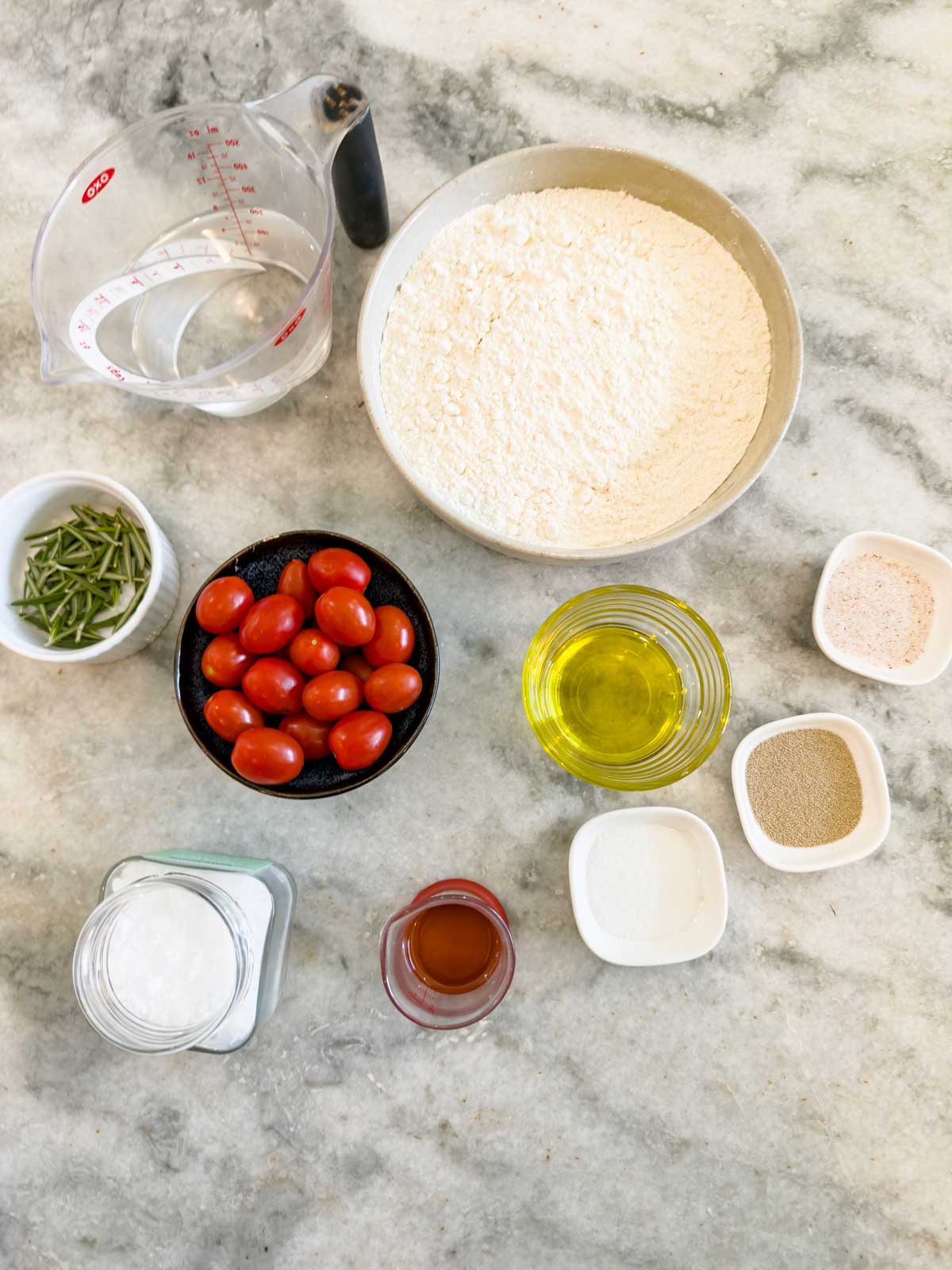 Top-down view of various ingredients on a marble counter for tomato focaccia bread, including flour, water, cherry tomatoes, olive oil, salt, yeast, sugar, fresh herbs, vinegar, and small bowls of seasonings.
