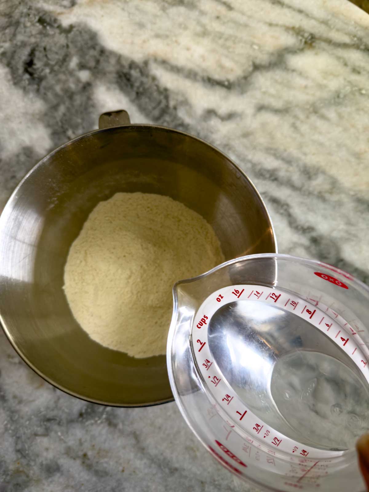 A metal mixing bowl with flour inside sits on a marble countertop. A hand holds a clear measuring cup filled with water above the bowl, ready to pour and begin making tomato focaccia bread.