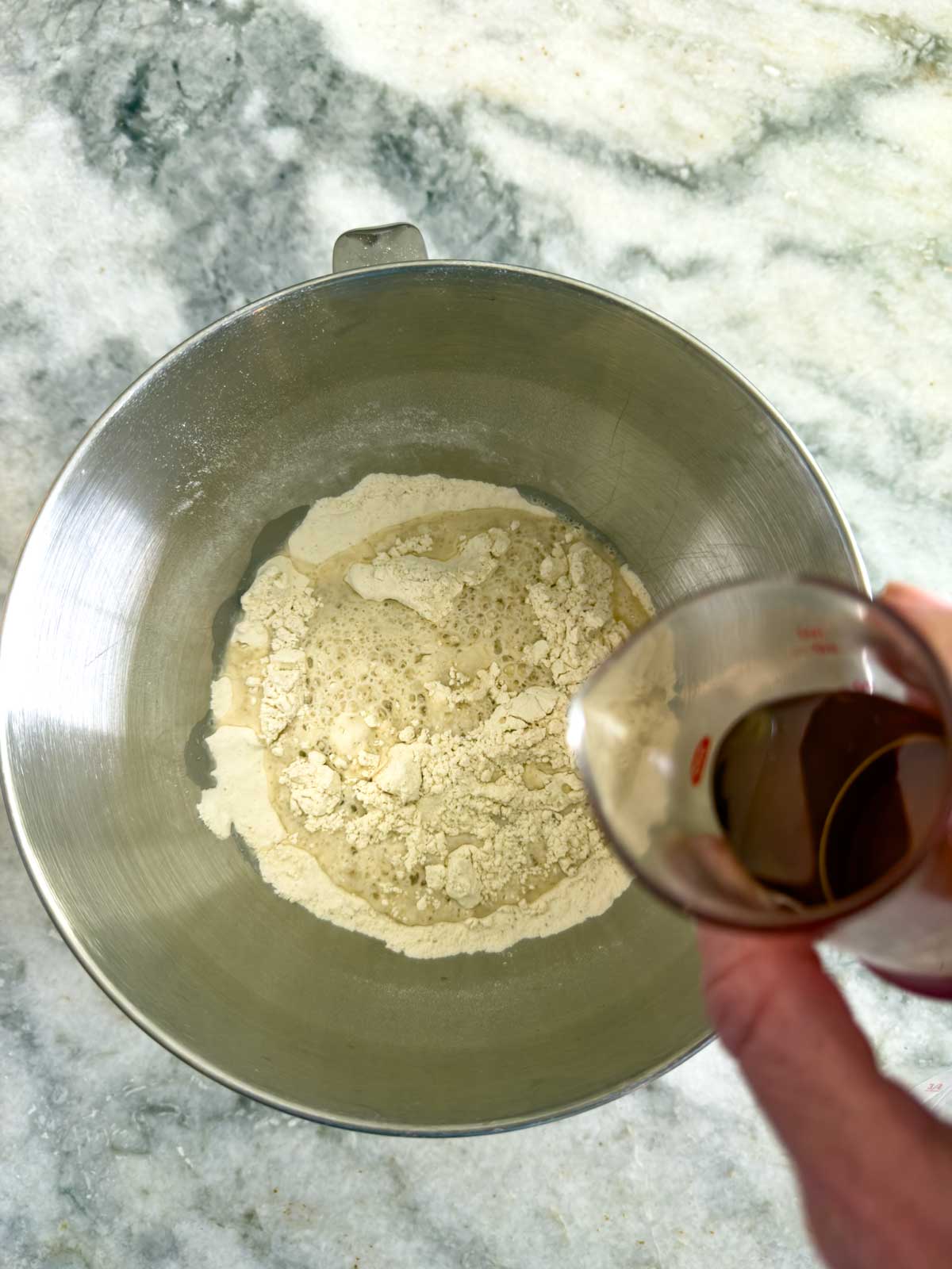 A hand pours liquid from a small measuring cup into a metal mixing bowl with flour and yeast, beginning the process of making tomato focaccia bread on a marble countertop.