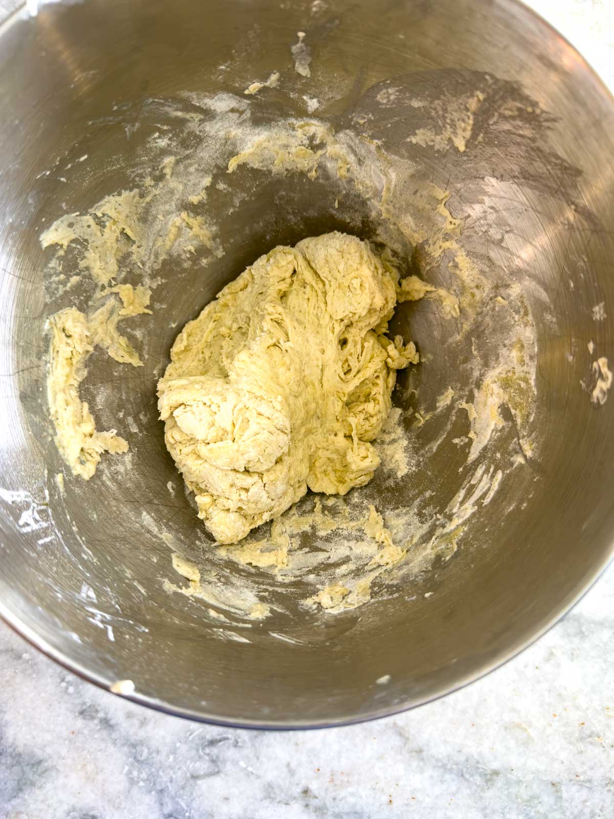 A ball of raw dough for tomato focaccia bread sits in the bottom of a large metal mixing bowl, with bits of flour and dough clinging to the sides. The bowl is placed on a light-colored countertop.