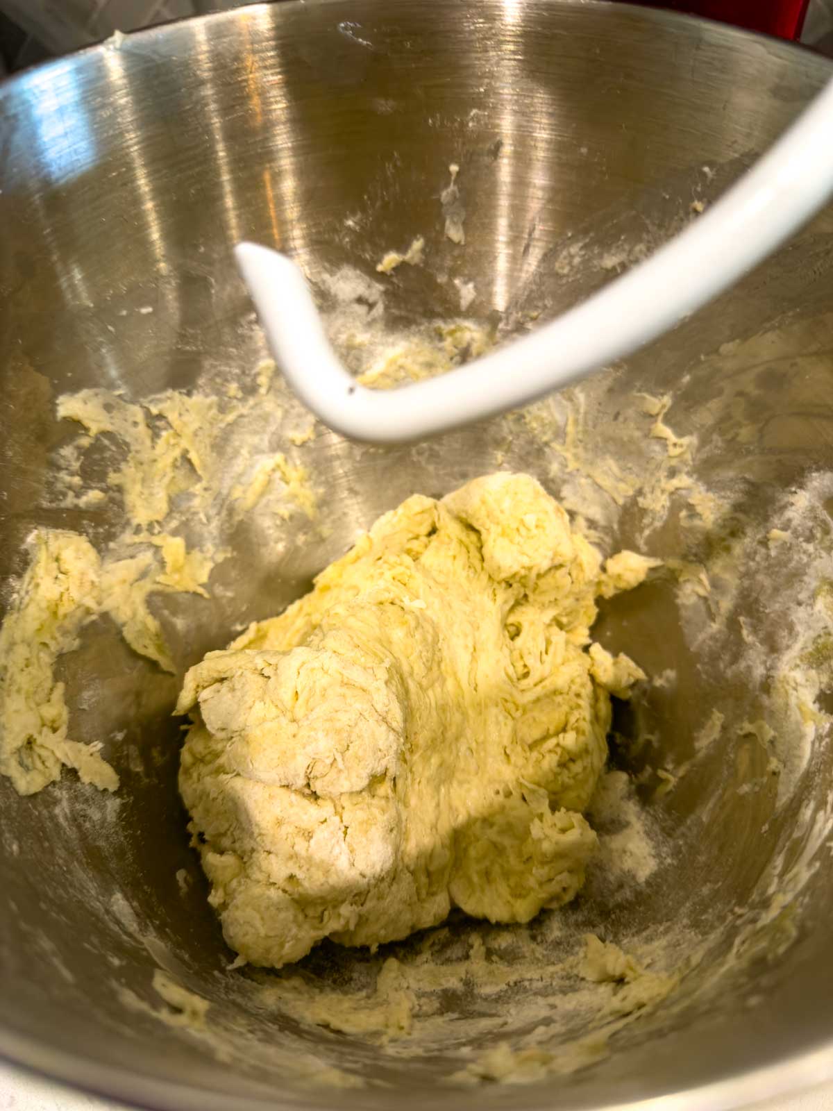 A metal mixing bowl with a dough hook attachment, containing a ball of pale, slightly sticky tomato focaccia bread dough in the process of being mixed.