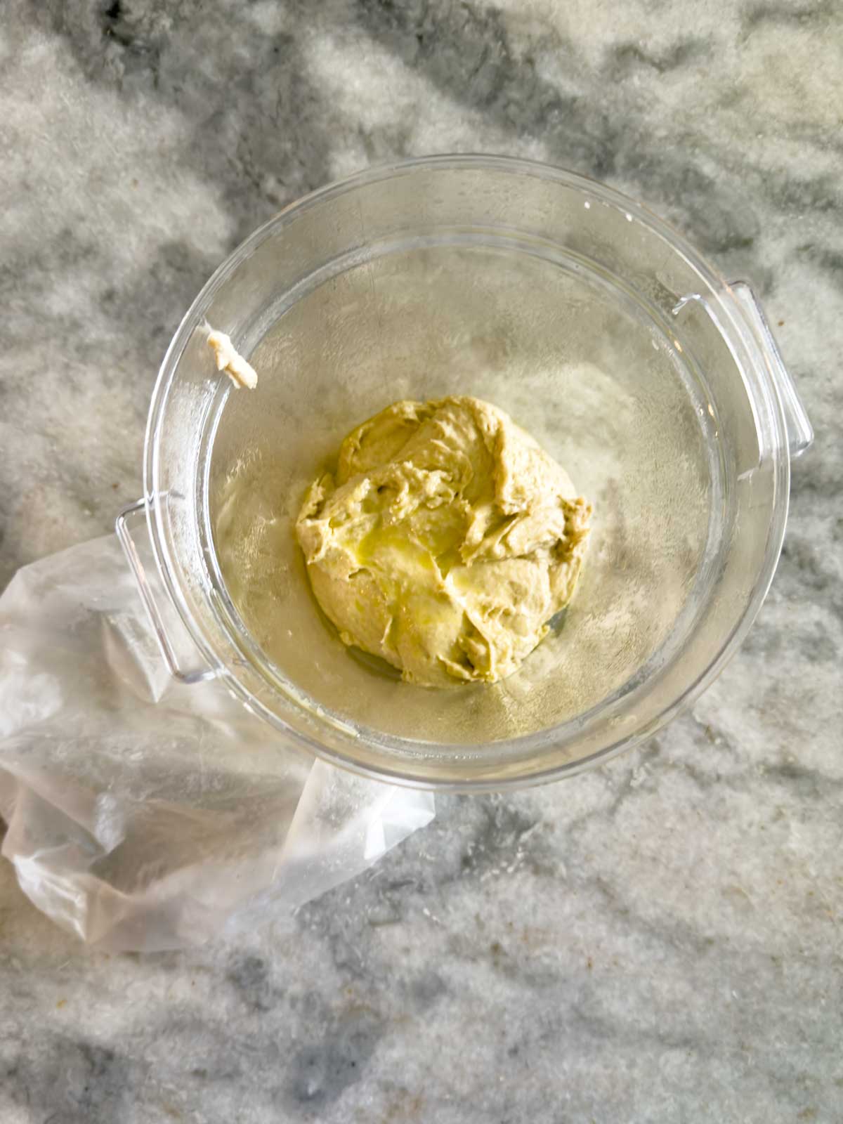 A ball of dough for tomato focaccia bread sits in the center of a glass bowl on a marble countertop, with a plastic covering partially off to the side.