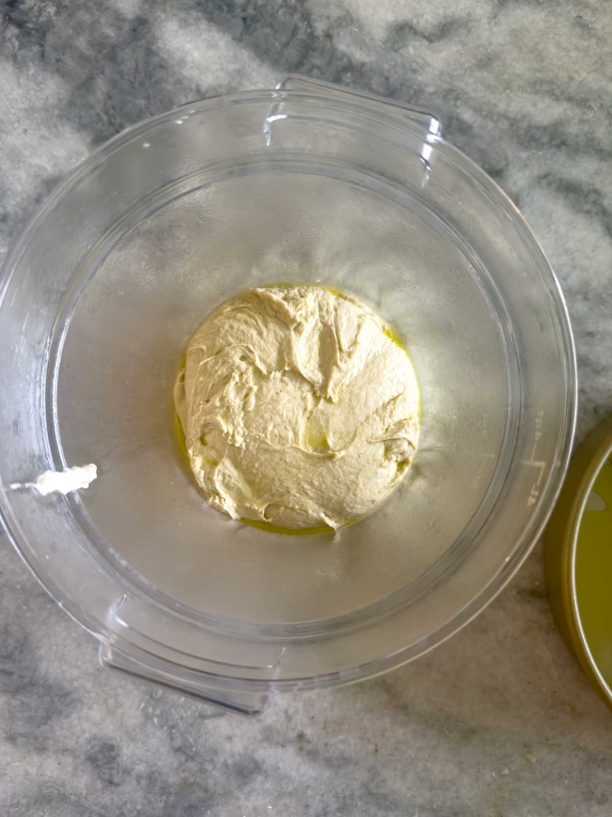 A food processor bowl holds a creamy, pale yellow mixture—likely fresh hummus—topped with a drizzle of olive oil. The lid rests beside it on a gray marbled countertop, perfect for enjoying with slices of tomato focaccia bread.