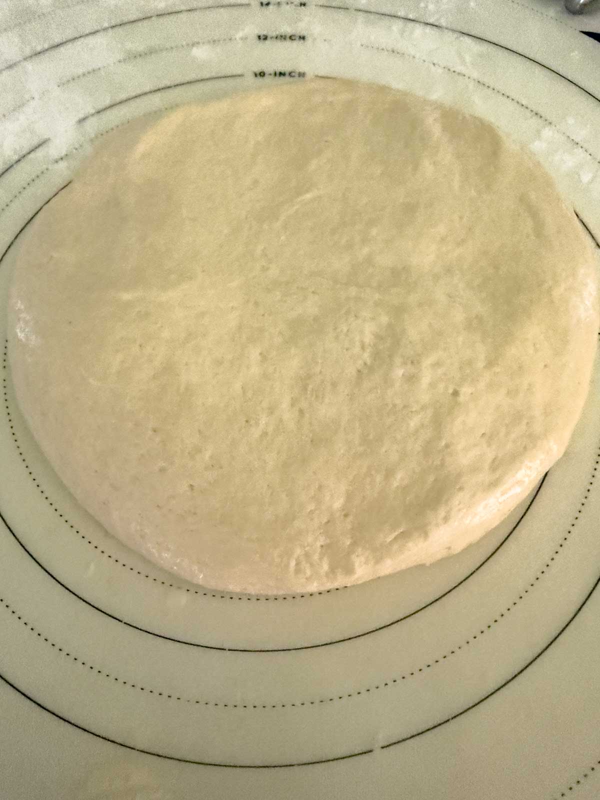 A round, risen tomato focaccia bread dough rests on a lightly floured surface with concentric measurement circles marked in inches, indicating preparation for baking or pizza making.
