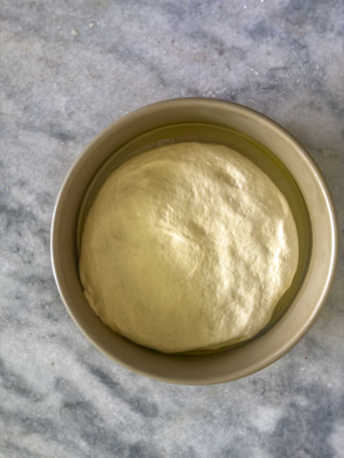 A round ball of dough for tomato focaccia bread rising in a metal bowl, set on a light gray marble surface.