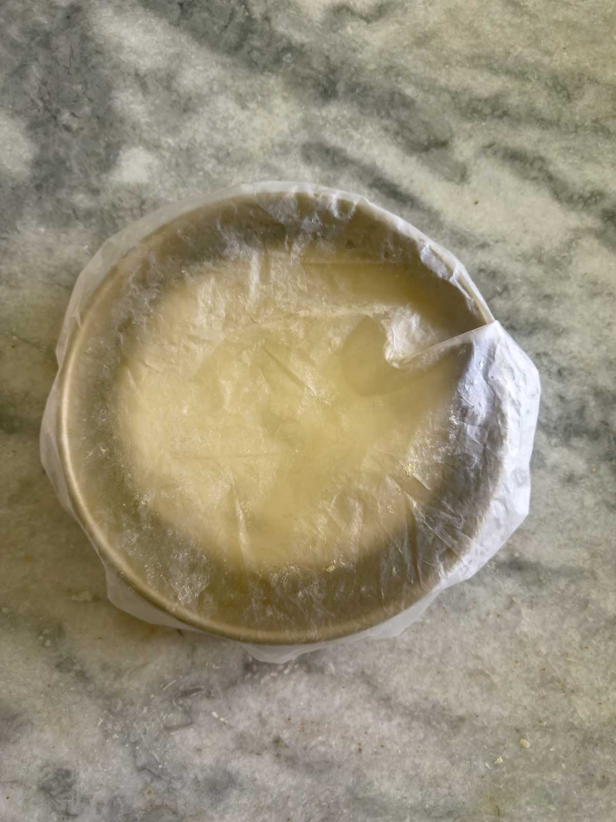 A small round bowl covered tightly with clear plastic wrap sits on a light gray marbled surface, ready for the tomato focaccia bread dough to rise.
