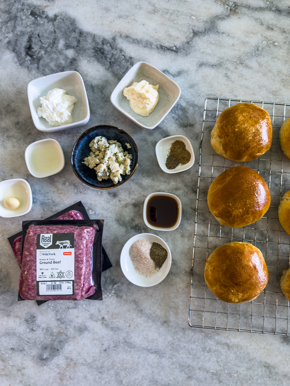 Ingredients for making burgers are arranged on a light stone surface, including brioche buns on a cooling rack, a package of ground beef, small bowls of sauces, seasonings, and crumbled cheese.