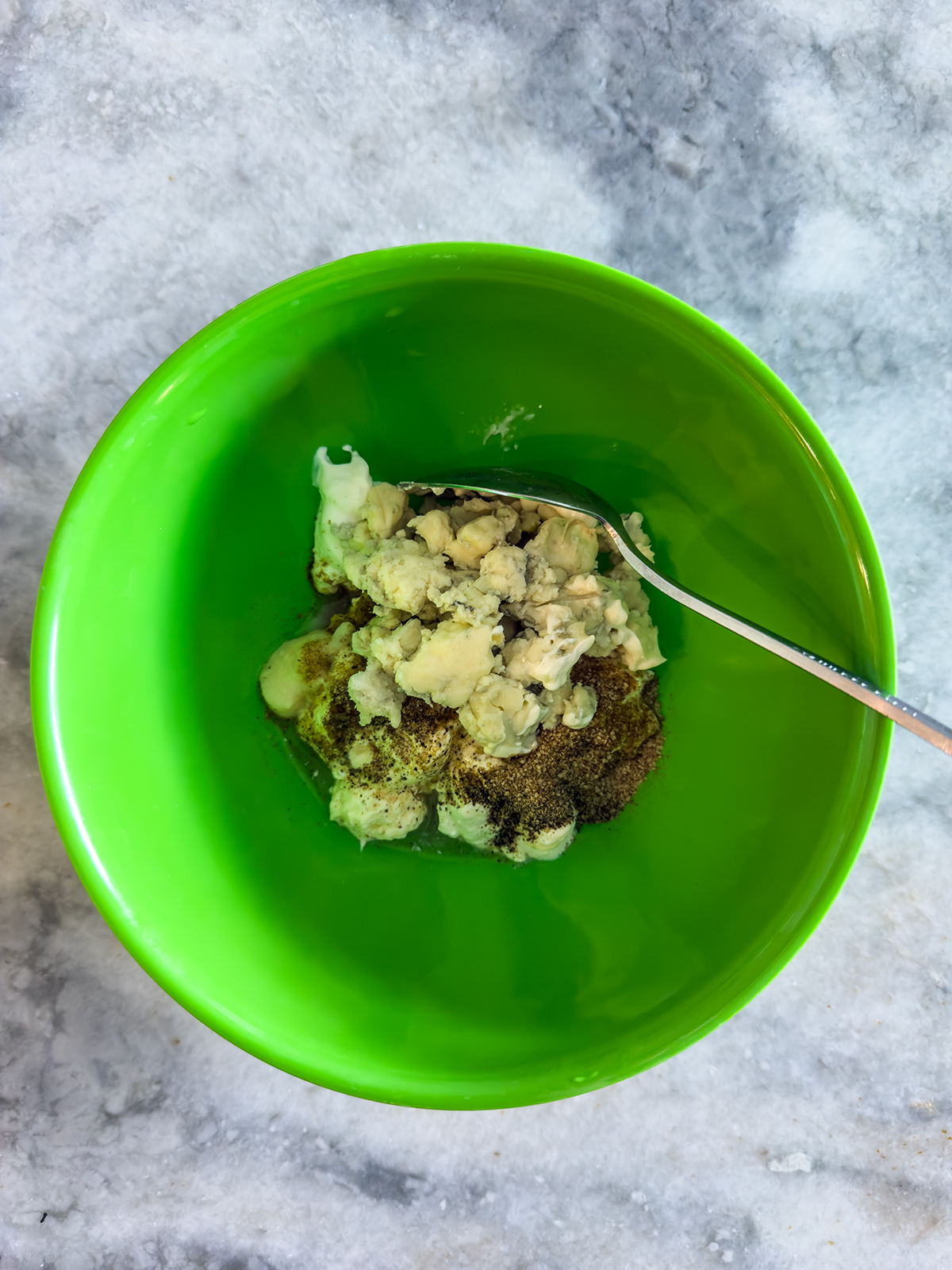 A green bowl containing a mixture of white creamy substance and brown spices, with a metal spoon resting inside, on a gray marble surface.