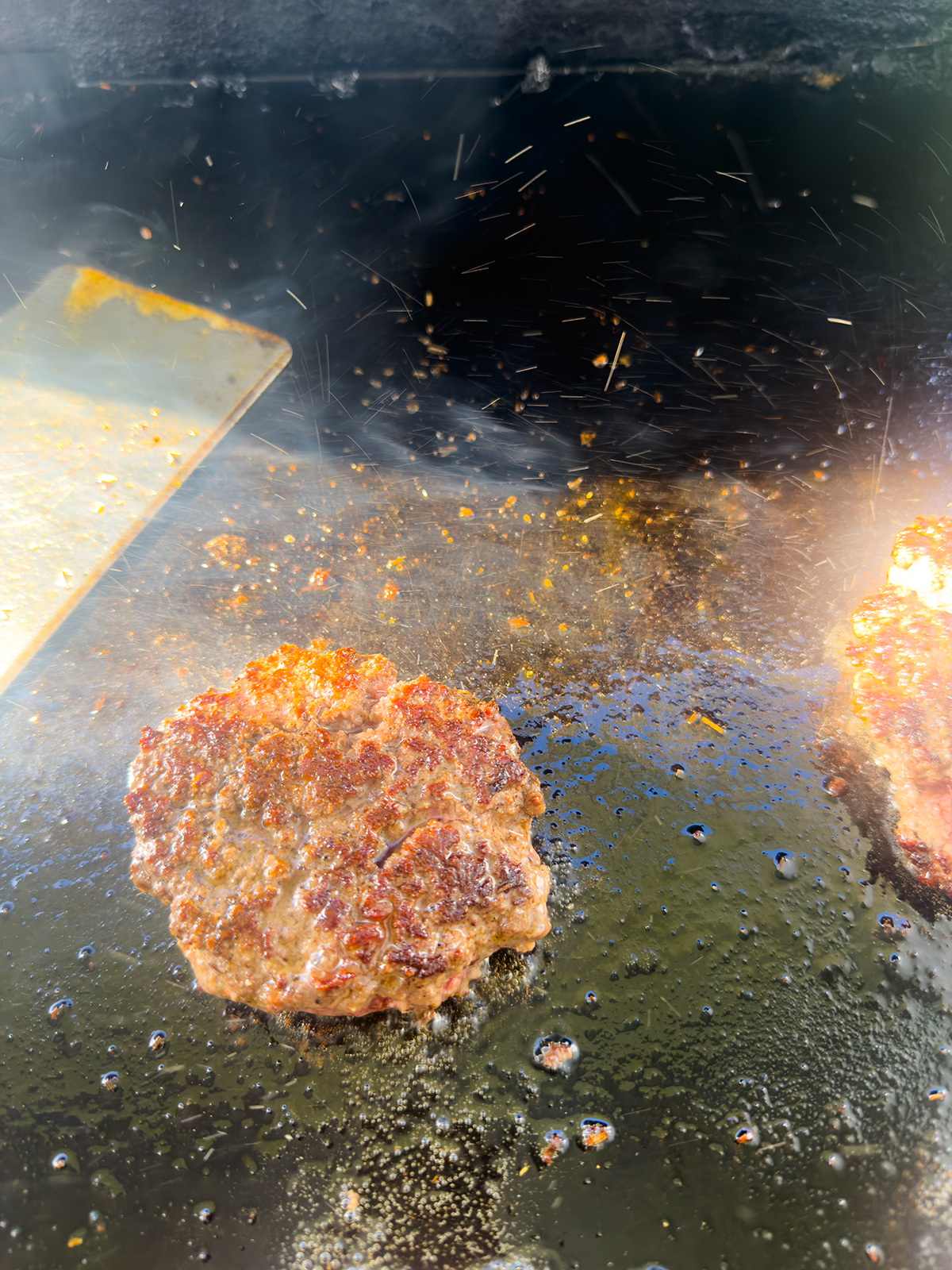 A close-up of hamburger patties sizzling on a greasy, hot griddle, with oil splatters and rising steam. A metal spatula is visible to the left and sparks or droplets light up the cooking surface.