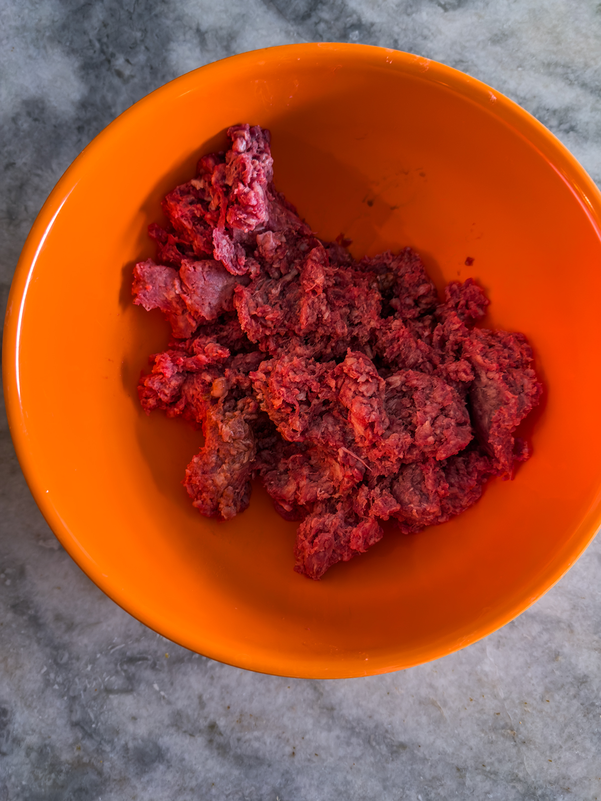Raw ground meat in clumps is placed in an orange bowl, set on a gray marble surface.