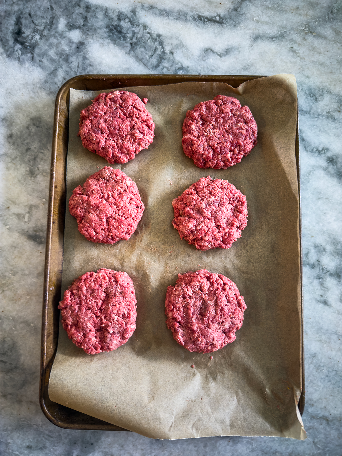 Six raw beef burger patties are arranged on a parchment-lined baking tray, placed on a mottled gray marble surface.
