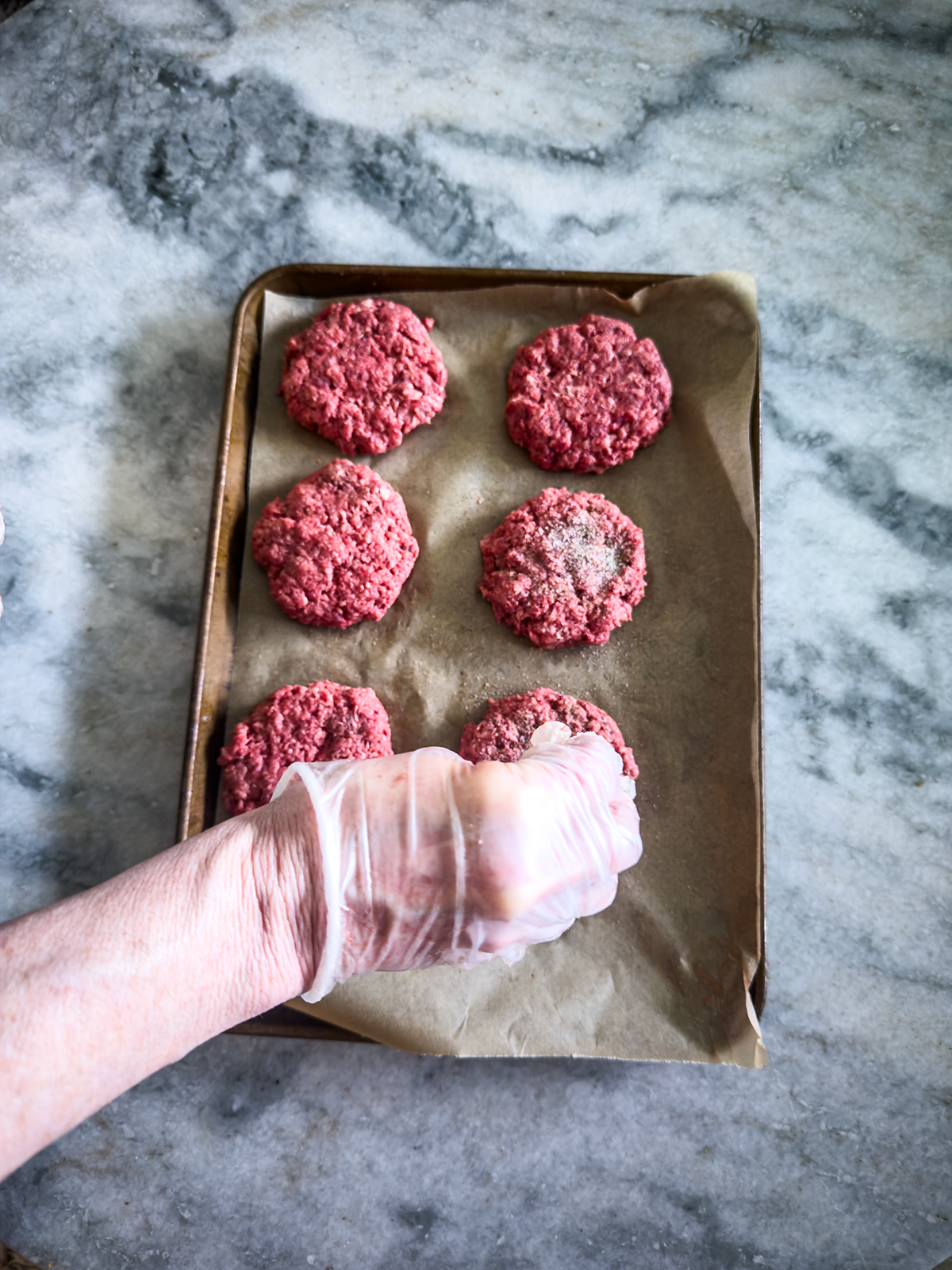A gloved hand places seasons raw hamburger patties on a baking sheet lined with parchment paper.