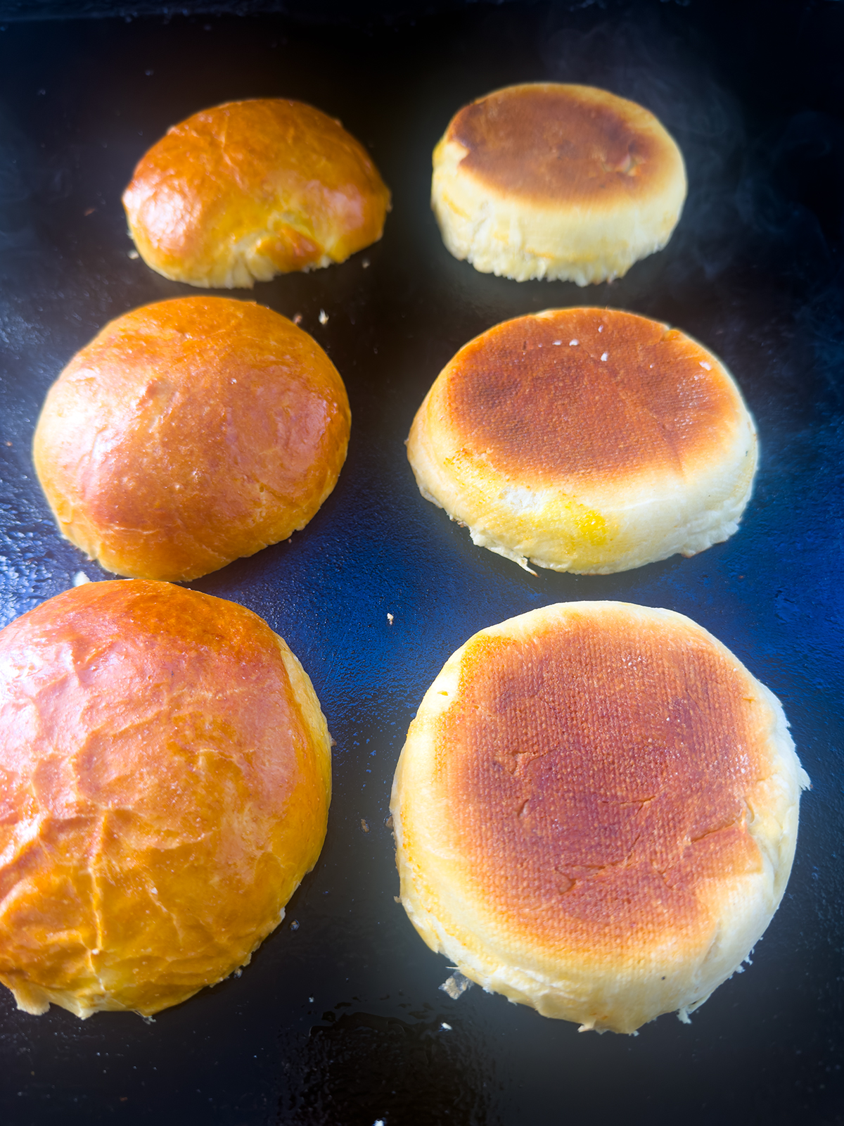 Six round bread rolls are being toasted on a skillet or griddle, with some showing a golden-brown, toasted underside while others display a shiny, un-toasted top.