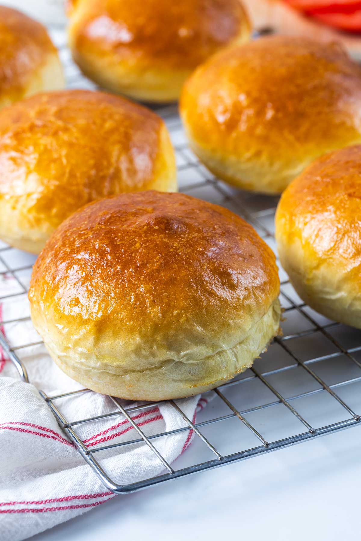 Golden brown, freshly baked hamburger buns are cooling on a metal wire rack, with a white kitchen towel featuring a red stripe partially visible in the foreground.